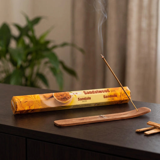 GR Sandelholz Räucherstäbchen: glimmendes Räucherstäbchen mit feinem Rauch im Holzhalter, dahinter die Sechseck-Packung, auf einem Sideboard am Abend