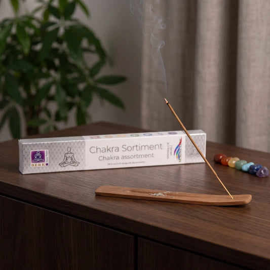 Chakra Line Sortimentspackung Holy Smokes: glimmendes Räucherstäbchen mit feinem Rauch im Holzhalter, dahinter die Packung, auf einem Sideboard am Abend
