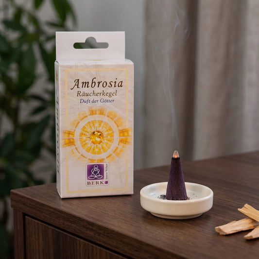 Räucherpyramiden Ambrosia von Berk: glimmender Räucherkegel mit feinem Rauch auf einem Keramiktellerchen, dahinter die Schachtel, auf einem Sideboard am Abend