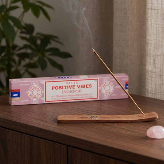 Satya Positive Vibes Räucherstäbchen: glimmendes Räucherstäbchen mit feinem Rauch im Holzhalter, dahinter die Packung, auf einem Sideboard am Abend