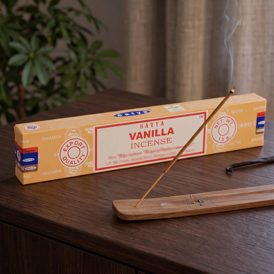 Satya Vanilla Räucherstäbchen: glimmendes Räucherstäbchen mit feinem Rauch im Holzhalter, dahinter die Packung, auf einem Sideboard am Abend