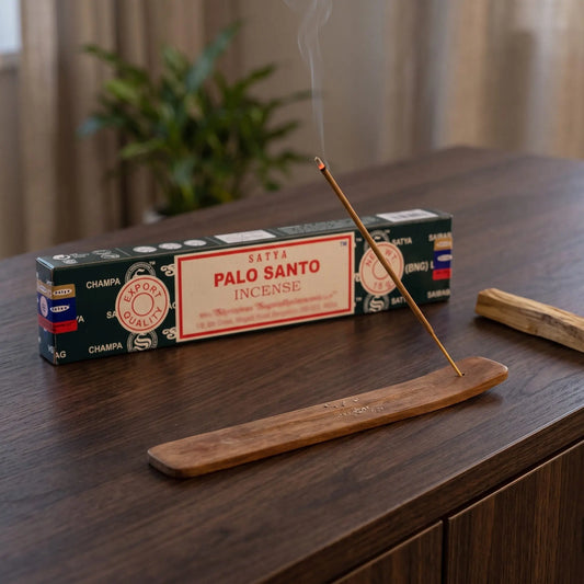 Satya Palo Santo Räucherstäbchen: glimmendes Räucherstäbchen mit feinem Rauch im Holzhalter, dahinter die Packung, auf einem Sideboard am Abend