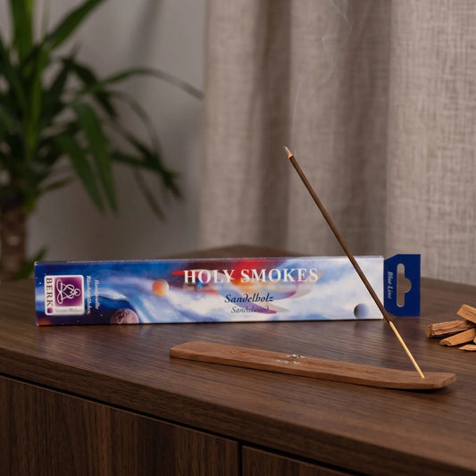 Sandelholz Räucherstäbchen Holy Smokes Blue Line: glimmendes Räucherstäbchen mit feinem Rauch im Holzhalter, dahinter die Packung, auf einem Sideboard am Abend