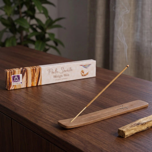 Räucherstäbchen Palo Santo von Berk: glimmendes Räucherstäbchen mit feinem Rauch im Holzhalter, dahinter die Packung, auf einem Sideboard am Abend