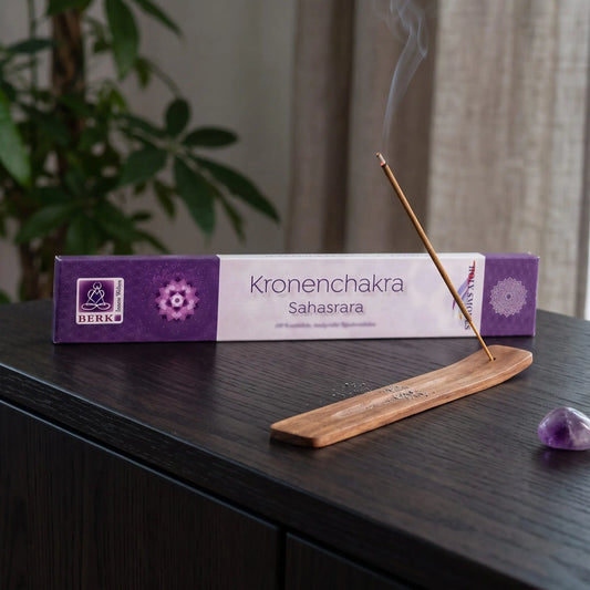 Kronenchakra Räucherstäbchen Holy Smokes: glimmendes Räucherstäbchen mit feinem Rauch im Holzhalter, dahinter die Packung, auf einem Sideboard am Abend