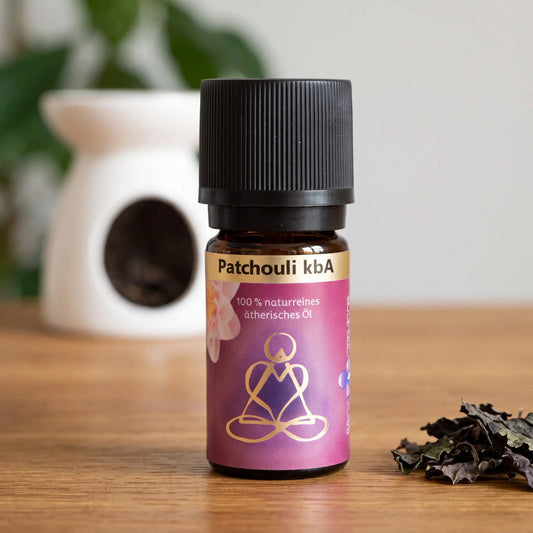 Ätherisches Öl Patchouli: Fläschchen auf einem Holz-Sideboard im Wohnzimmer, im Hintergrund eine weiße Duftlampe, daneben Patchouliblätter