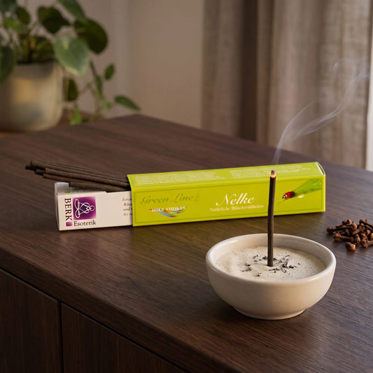 Nelke Räucherstäbchen naturrein, Holy Smokes Green Line: glimmendes Räucherstäbchen in einer Keramikschale mit Sand, dahinter die geöffnete Packung, auf einem Sideboard am Abend