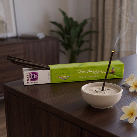 Champa-Blüten Räucherstäbchen naturrein, Holy Smokes Green Line: glimmendes Räucherstäbchen in einer Keramikschale mit Sand, dahinter die geöffnete Packung, auf einem Sideboard am Abend
