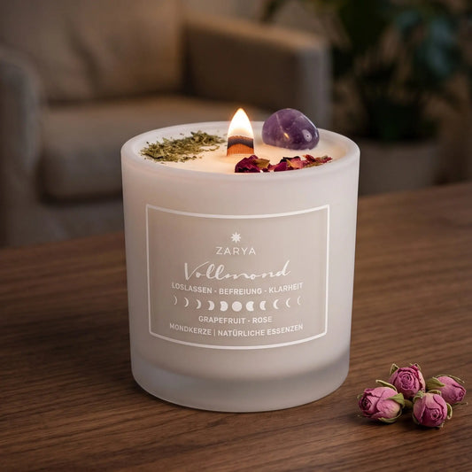 Zeremonie Kerze Vollmond mit Amethyst, Duft Grapefruit und Rose: brennende Kerze mit kleiner Flamme am Abend auf einer dunklen Holzplatte im Wohnzimmer, daneben ein paar getrocknete Rosenknospen – Spirit of Gaia