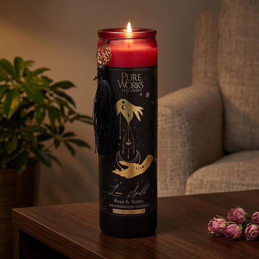 Manifestationskerze Love Spell (Rose & Pfingstrose): brennende Glaskerze auf einem dunklen Holztisch im Wohnzimmer am Abend, daneben getrocknete Rosenknospen