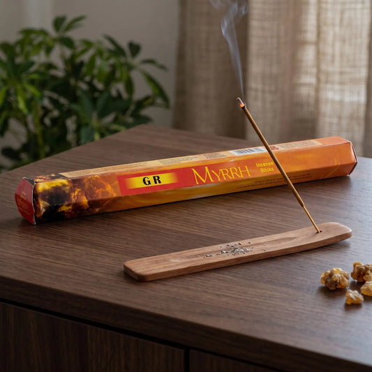 GR Myrrhe Räucherstäbchen: glimmendes Räucherstäbchen mit feinem Rauch im Holzhalter, dahinter die Sechseck-Packung, auf einem Sideboard am Abend
