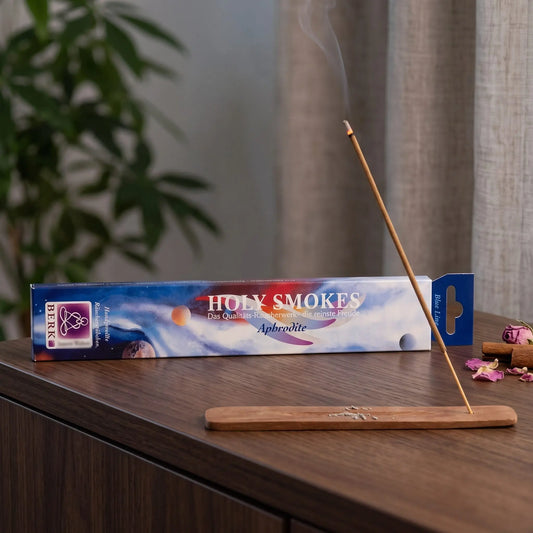 Aphrodite Räucherstäbchen Blue Line: glimmendes Räucherstäbchen mit feinem Rauch im Holzhalter, dahinter die Packung, auf einem Sideboard am Abend