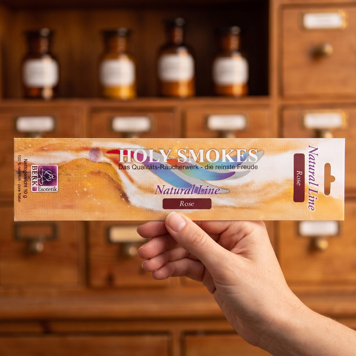 Eine Hand hält die flache Packung Rose Natural Line von Holy Smokes, etwa 24 cm lang, vor einem Holzschrank mit Schubladen