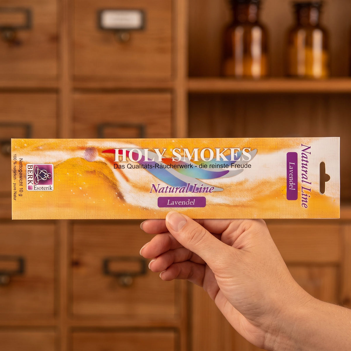 Eine Hand hält die flache Packung Lavendel Natural Line von Holy Smokes, etwa 24 cm lang, vor einem Holzschrank mit Schubladen