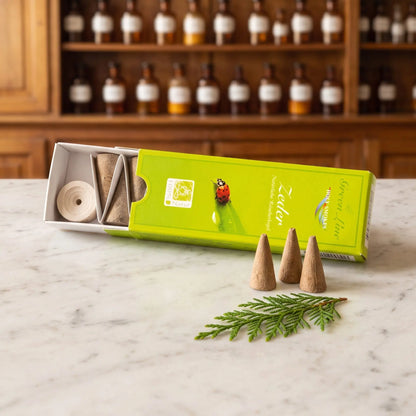Zeder Räucherkegel naturrein, Holy Smokes Green Line: geöffnete Packung auf einer Marmortheke vor einem Apothekerschrank, davor lose Räucherkegel und einem Zedernzweig – Spirit of Gaia