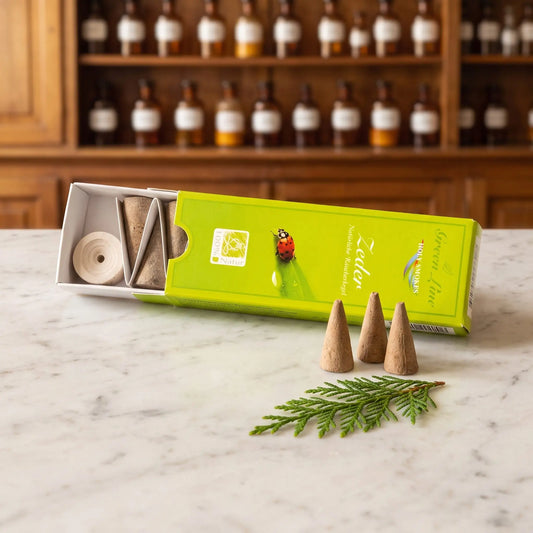 Zeder Räucherkegel naturrein, Holy Smokes Green Line: geöffnete Packung auf einer Marmortheke vor einem Apothekerschrank, davor lose Räucherkegel und einem Zedernzweig – Spirit of Gaia