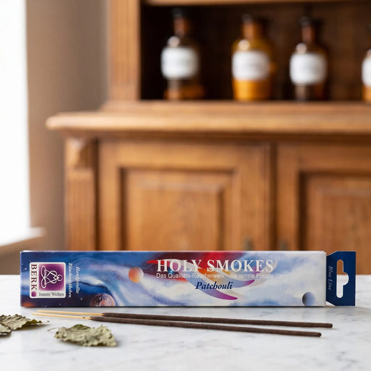 Patchouli Räucherstäbchen Holy Smokes Blue Line: Packung auf einer Marmortheke vor einem Apothekerschrank, davor zwei lose Räucherstäbchen und getrocknete Patchouliblätter – Spirit of Gaia