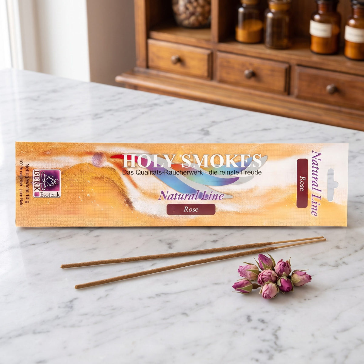 Rose Räucherstäbchen der Holy Smokes Natural Line von Berk: flache Packung auf einer Marmortheke vor einem alten Holzschrank mit Schubladen und Bernsteinglasflaschen, davor zwei lose Stäbchen und getrocknete Rosenknospen