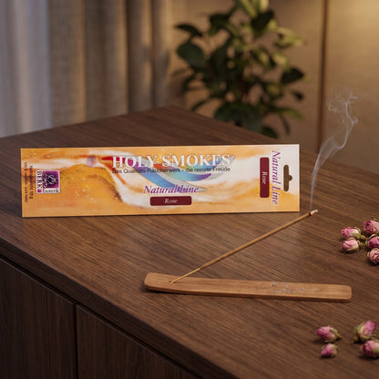 Rose Räucherstäbchen Natural Line: glimmendes Stäbchen im Holzhalter auf einem dunklen Sideboard, dahinter die Packung, am Abend