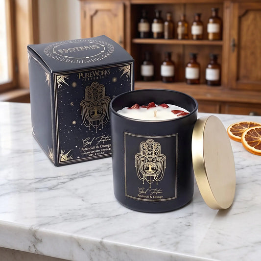 Manifestationskerze Good Fortune mit rotem Jaspis (Patchouli & Orange) von Pure Works: große Kerze im schwarzen Glas mit Golddeckel und schwarze Schachtel mit Goldprägung auf Marmortheke vor Apothekerschrank, daneben getrocknete Orangenscheiben – Spirit of Gaia