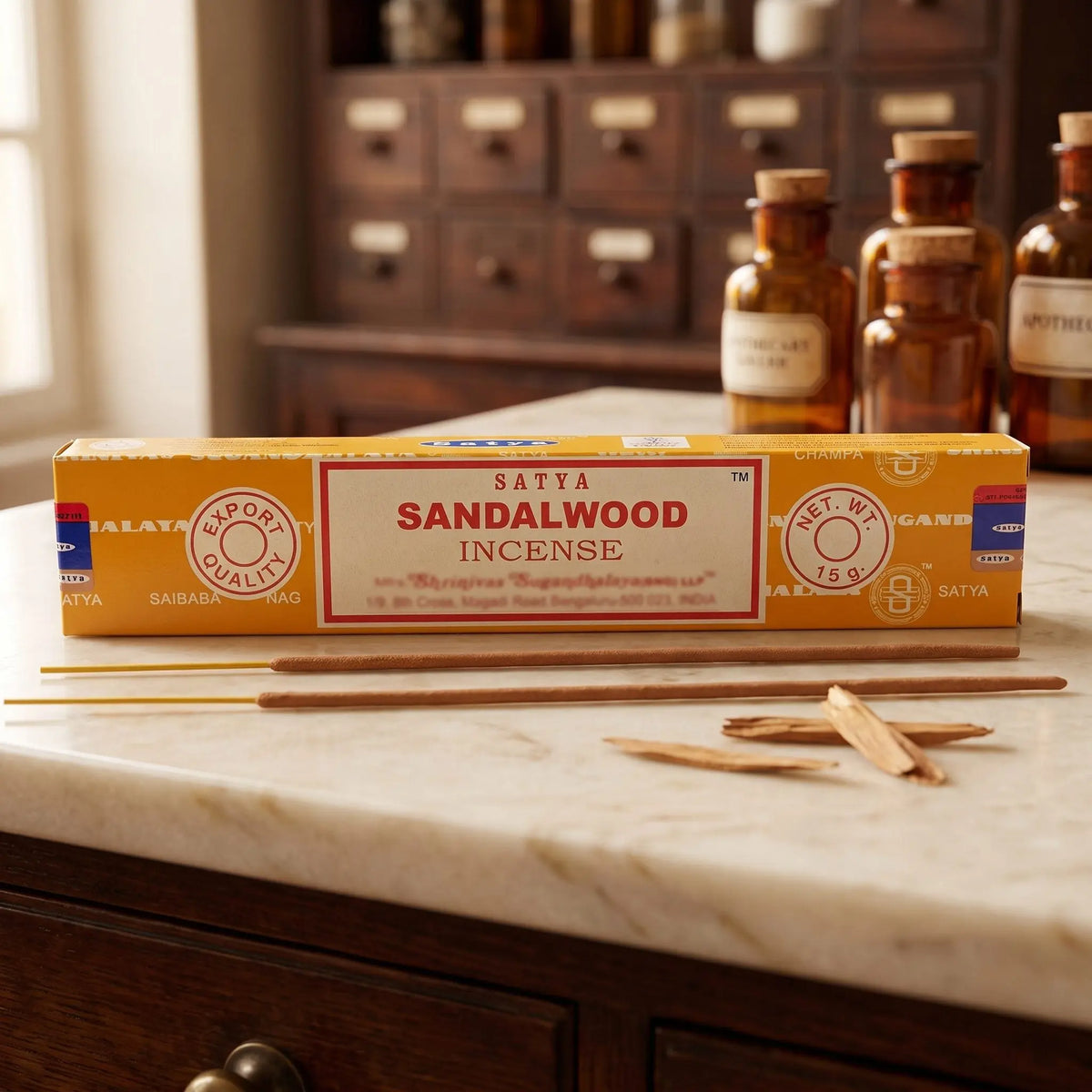 Satya Sandalwood Räucherstäbchen: 15-g-Packung auf einer Marmortheke vor einem Apothekerschrank, daneben zwei lose Räucherstäbchen – Spirit of Gaia