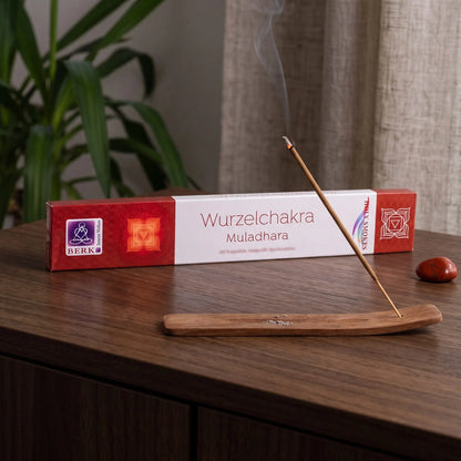 Wurzelchakra Räucherstäbchen Holy Smokes: glimmendes Räucherstäbchen mit feinem Rauch im Holzhalter, dahinter die Packung, auf einem Sideboard am Abend