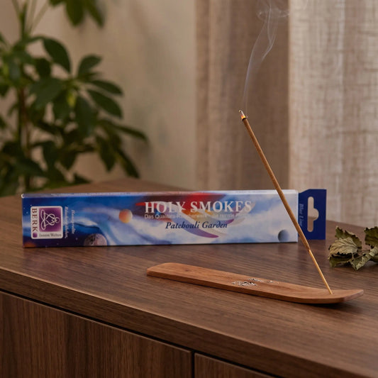 Patchouli Garden Räucherstäbchen Blue Line: glimmendes Räucherstäbchen mit feinem Rauch im Holzhalter, dahinter die Packung, auf einem Sideboard am Abend