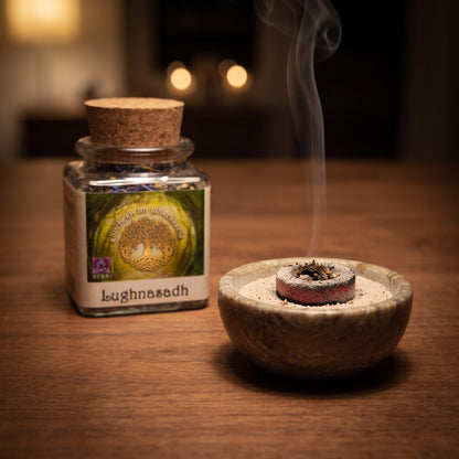 Lughnasadh, Jahreskreisfeste-Räuchermischung von Berk, 50 ml im Korkglas: glimmende Räucherkohle mit einer Prise Räucherwerk in einer Specksteinschale, dahinter das Korkglas, feiner Rauch steigt auf einer dunklen Holzplatte am Abend auf – Spirit of Gaia