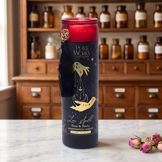 Manifestationskerze Love Spell (Rose & Pfingstrose) von Pure Works: hohe Glaskerze mit rotem Glasrand, schwarzer Quaste und Goldanhänger auf Marmortheke vor Apothekerschrank, daneben getrocknete Rosenknospen – Spirit of Gaia