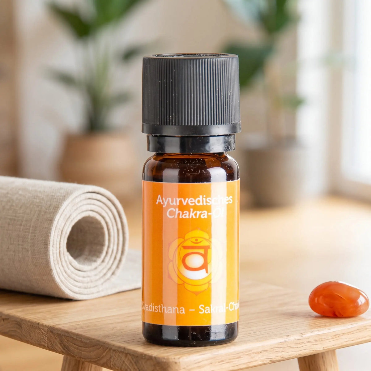 Ayurvedisches Chakra-Öl Svadisthana (Sakral-Chakra): 10-ml-Fläschchen auf einem Holzhocker neben einer gerollten Yogamatte, daneben Karneol