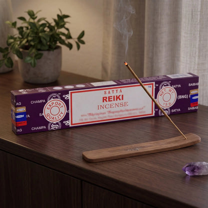 Satya Reiki Räucherstäbchen: glimmendes Räucherstäbchen mit feinem Rauch im Holzhalter, dahinter die Packung, auf einem Sideboard am Abend