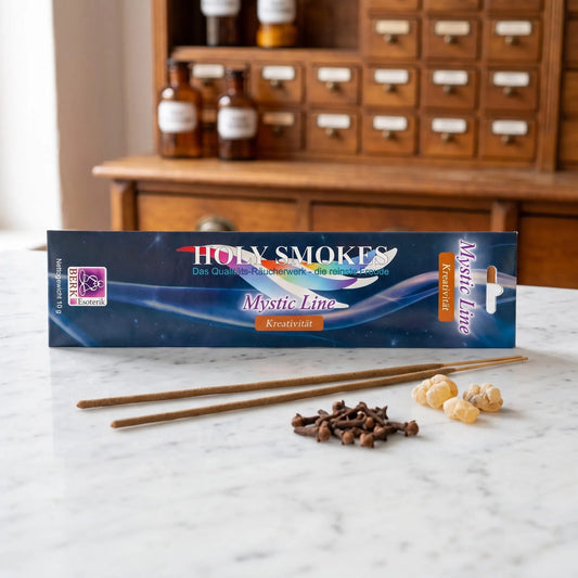 Kreativität Räucherstäbchen Holy Smokes Mystic Line: Packung auf einer Marmortheke vor einem Apothekerschrank, davor zwei lose Räucherstäbchen, Nelken und Weihrauchharz – Spirit of Gaia