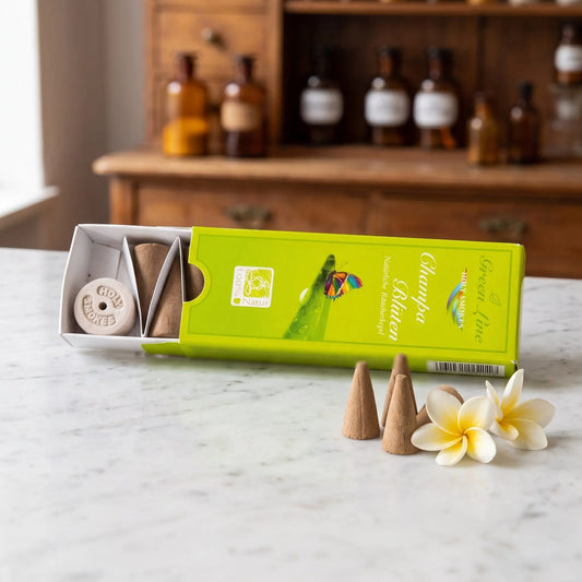 Champa-Blüten Räucherkegel naturrein, Holy Smokes Green Line: geöffnete Packung auf einer Marmortheke vor einem Apothekerschrank, davor lose Räucherkegel und Champa-Blüten – Spirit of Gaia