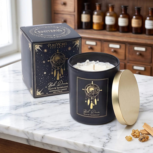Manifestationskerze Boho Dreams mit Bergkristall (Myrrhe & Sandelholz) von Pure Works: große Kerze im schwarzen Glas mit Golddeckel und schwarze Schachtel mit Goldprägung auf Marmortheke vor Apothekerschrank, daneben Myrrheharz und Sandelholzspäne – Spirit of Gaia