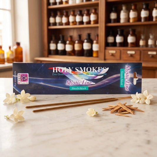 Sinnlichkeit Räucherstäbchen Holy Smokes Mystic Line: Packung auf einer Marmortheke vor einem Apothekerschrank, davor zwei lose Räucherstäbchen, weiße Blüten und Sandelholz – Spirit of Gaia
