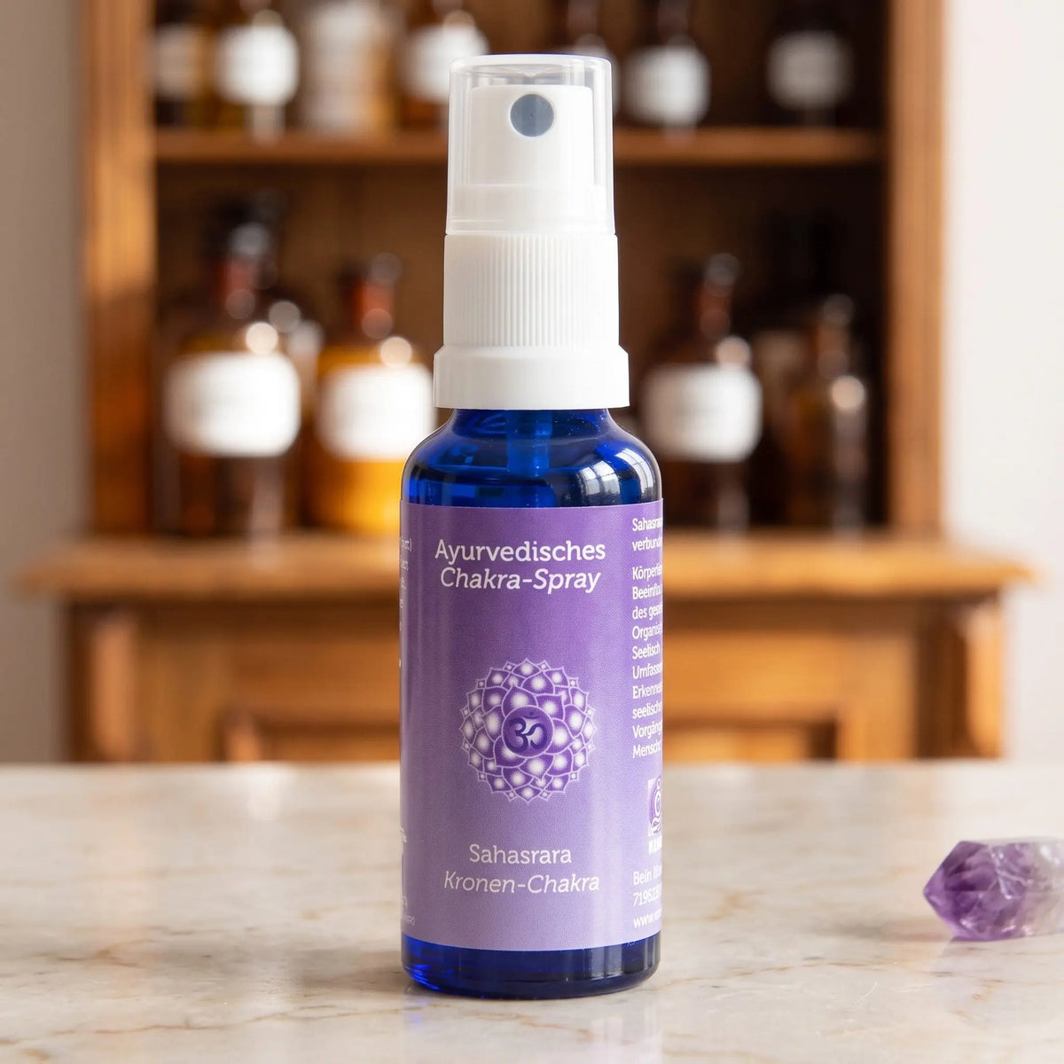Ayurvedisches Chakra-Spray Sahasrara (Kronen-Chakra): 30-ml-Sprühflasche aus blauem Glas auf Marmortheke vor Apothekerschrank, daneben Amethyst – Spirit of Gaia