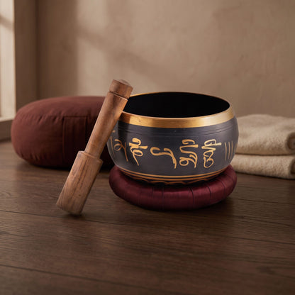 Schwarze Klangschale OM mit Mantra auf dunkelroter Unterlage und Klangstab auf dem Holzboden eines Meditationsraums