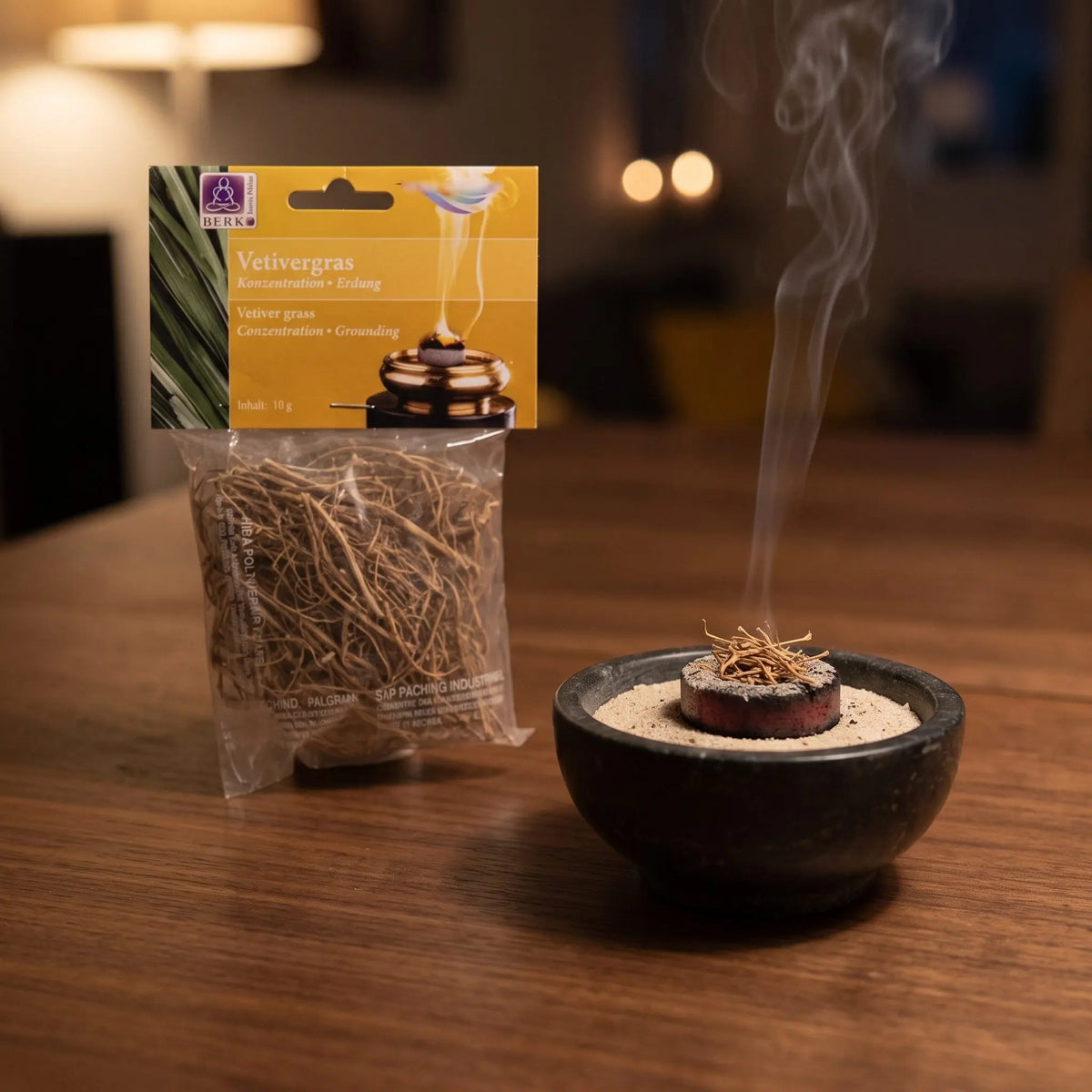 Vetivergras (Wurzel), sortenreines Räucherwerk von Berk, 10 g: ein Büschel Vetiverwurzeln glimmt auf der Räucherkohle, dahinter die Tüte, feiner Rauch steigt auf einer dunklen Holzplatte am Abend auf – Spirit of Gaia