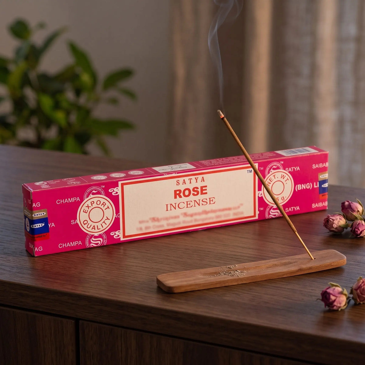 Satya Rose Räucherstäbchen: glimmendes Räucherstäbchen mit feinem Rauch im Holzhalter, dahinter die Packung, auf einem Sideboard am Abend
