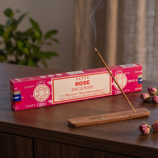 Satya Rose Räucherstäbchen: glimmendes Räucherstäbchen mit feinem Rauch im Holzhalter, dahinter die Packung, auf einem Sideboard am Abend