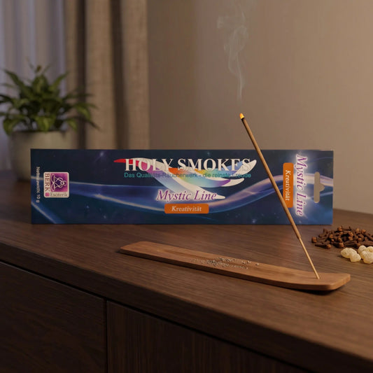 Kreativität Räucherstäbchen Holy Smokes Mystic Line: glimmendes Räucherstäbchen mit feinem Rauch im Holzhalter, dahinter die Packung, auf einem Sideboard am Abend