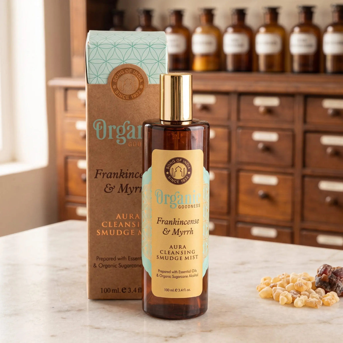 Aura Reinigungsspray Weihrauch und Myrrhe von Organic Goodness: braune Sprühflasche mit goldener Kappe neben der Kraftpapier-Schachtel auf einer Marmortheke vor einem Apothekerschrank
