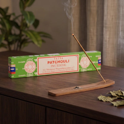 Satya Patchouli Räucherstäbchen: glimmendes Räucherstäbchen mit feinem Rauch im Holzhalter, dahinter die Packung, auf einem Sideboard am Abend