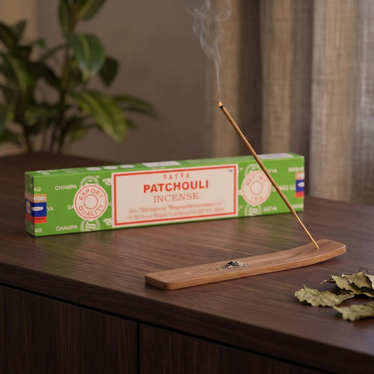 Satya Patchouli Räucherstäbchen: glimmendes Räucherstäbchen mit feinem Rauch im Holzhalter, dahinter die Packung, auf einem Sideboard am Abend