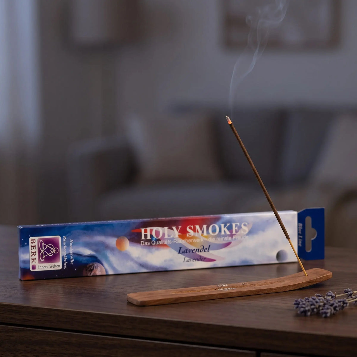 Lavendel Räucherstäbchen Holy Smokes Blue Line: glimmendes Räucherstäbchen mit feinem Rauch im Holzhalter, dahinter die Packung, auf einem Sideboard am Abend