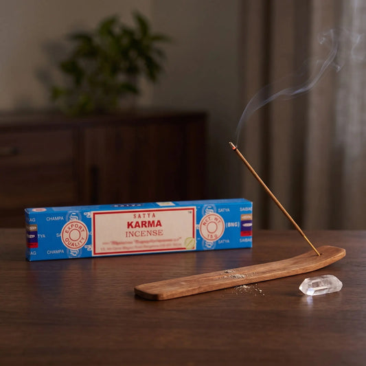Satya Karma Räucherstäbchen: glimmendes Räucherstäbchen mit feinem Rauch im Holzhalter, dahinter die Packung, auf einem Sideboard am Abend