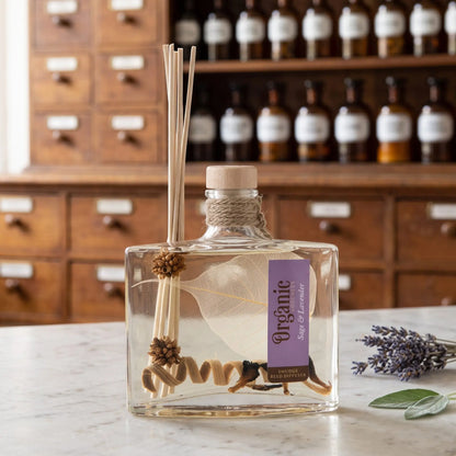 Reed Diffuser Salbei und Lavendel von Organic Goodness: eckige Glasflasche mit Holzstopfen, Jutekordel und Schilfrohrstäbchen auf einer Marmortheke vor einem Apothekerschrank