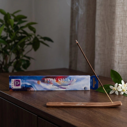 Orangenblüten Räucherstäbchen Holy Smokes Blue Line: glimmendes Räucherstäbchen mit feinem Rauch im Holzhalter, dahinter die Packung, auf einem Sideboard am Abend