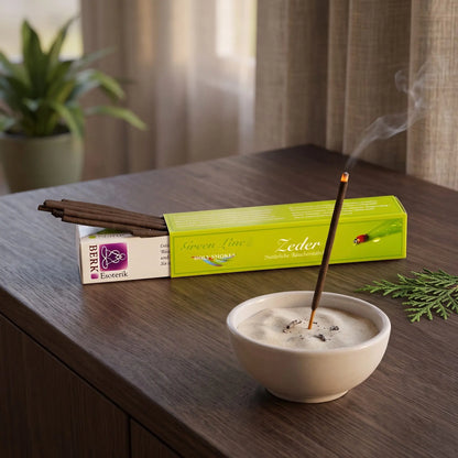Zeder Räucherstäbchen naturrein, Holy Smokes Green Line: glimmendes Räucherstäbchen in einer Keramikschale mit Sand, dahinter die geöffnete Packung, auf einem Sideboard am Abend