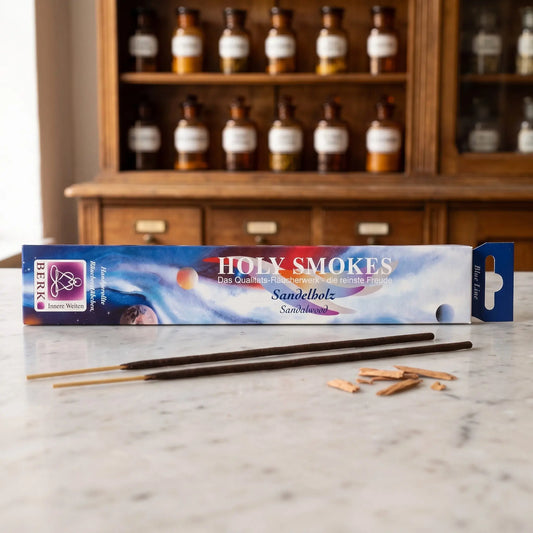 Sandelholz Räucherstäbchen Holy Smokes Blue Line: Packung auf einer Marmortheke vor einem Apothekerschrank, davor zwei lose Räucherstäbchen und Sandelholzspäne – Spirit of Gaia